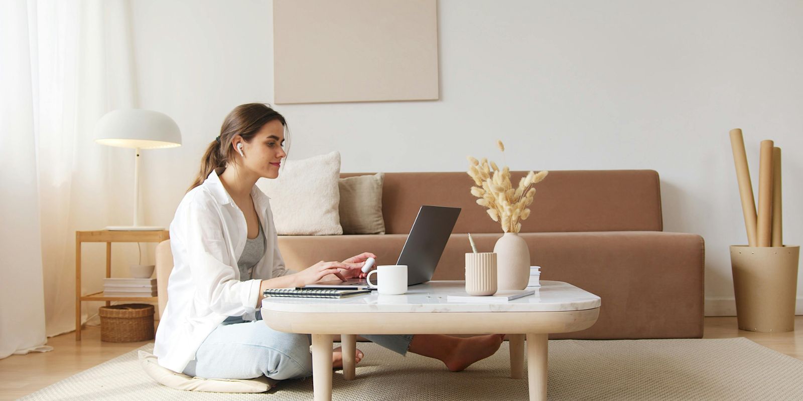 A focused individual is embracing the modern work-from-home lifestyle. Seated comfortably on the floor, they’re fully engaged with their laptop on a minimalistic coffee table. The space is bathed in natural light, creating a serene and productive ambiance perfect for concentrated tasks or online learning. The decor, with its muted tones and natural textures, suggests a penchant for contemporary, clutter-free living