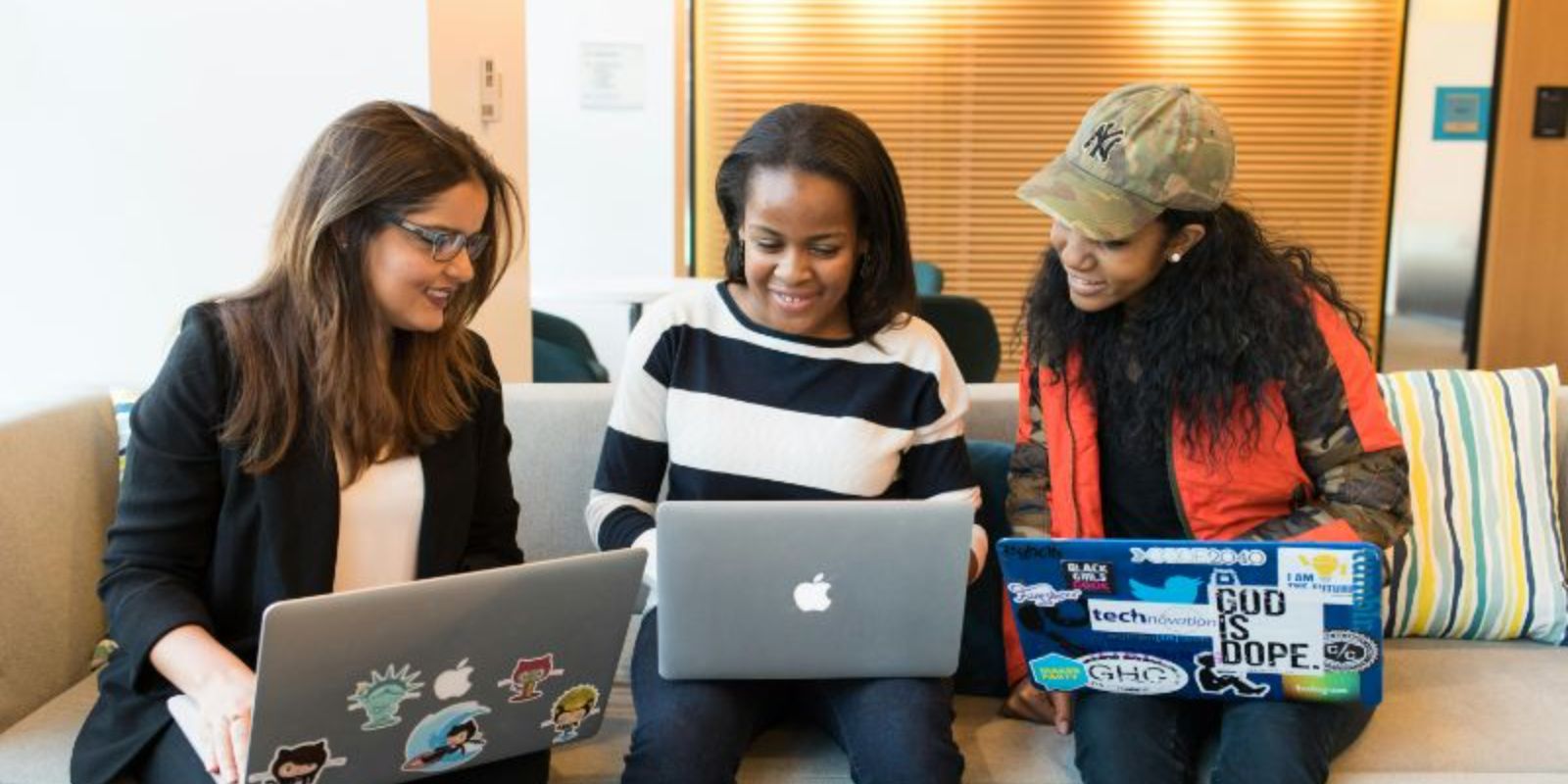 Three ladies sitting while looking at each other's laptops.
