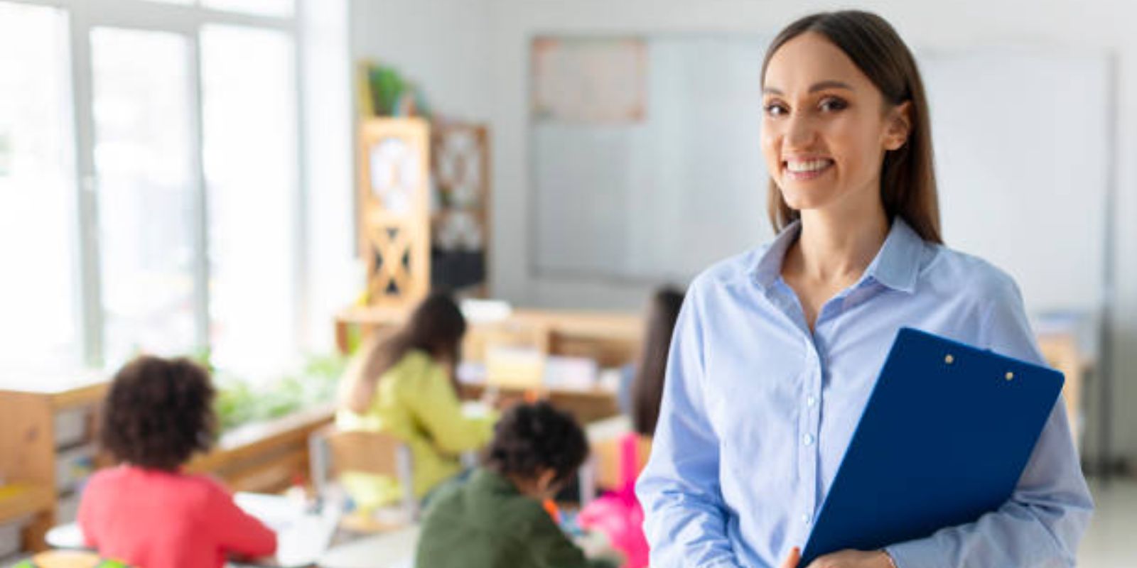 Portrait of cheerful female teacher standing in classroom during lesson with pupils studying on background