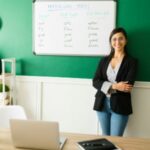 A young teacher leaning on the board while smiling.